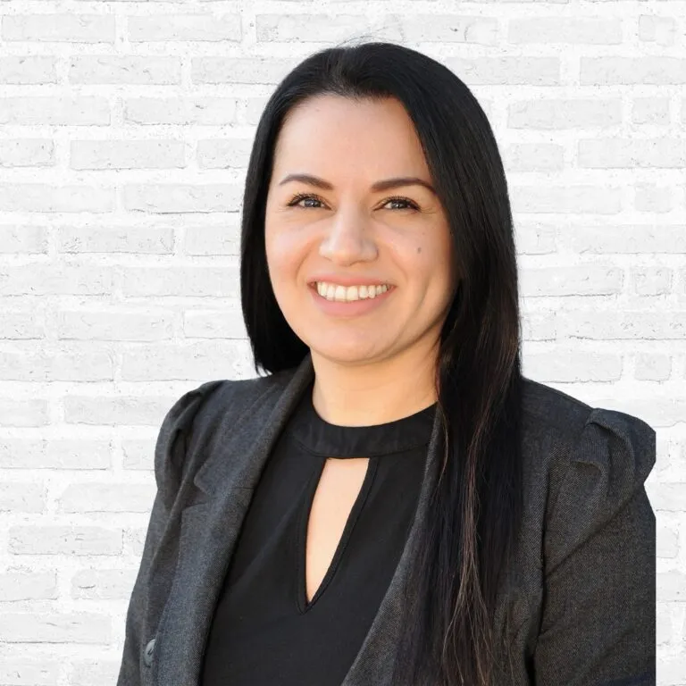 Smiling woman with long dark hair in a professional gray blazer stands against a white brick background.