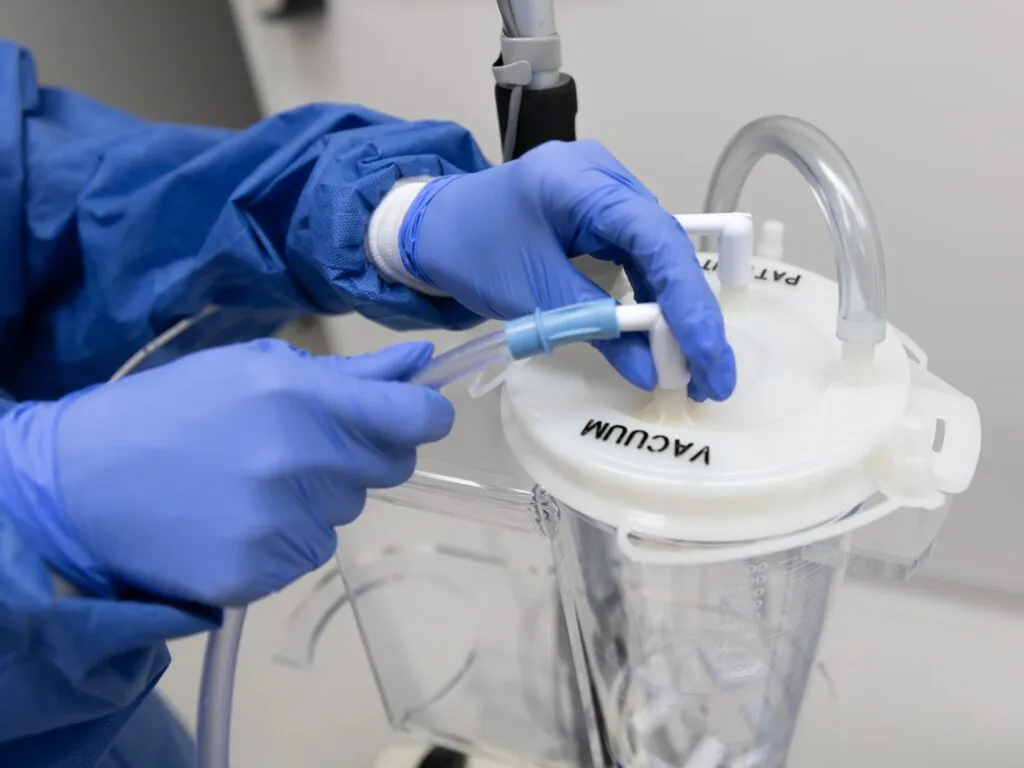 Gloved hands connecting surgical tubing to a vacuum canister in a medical setting.