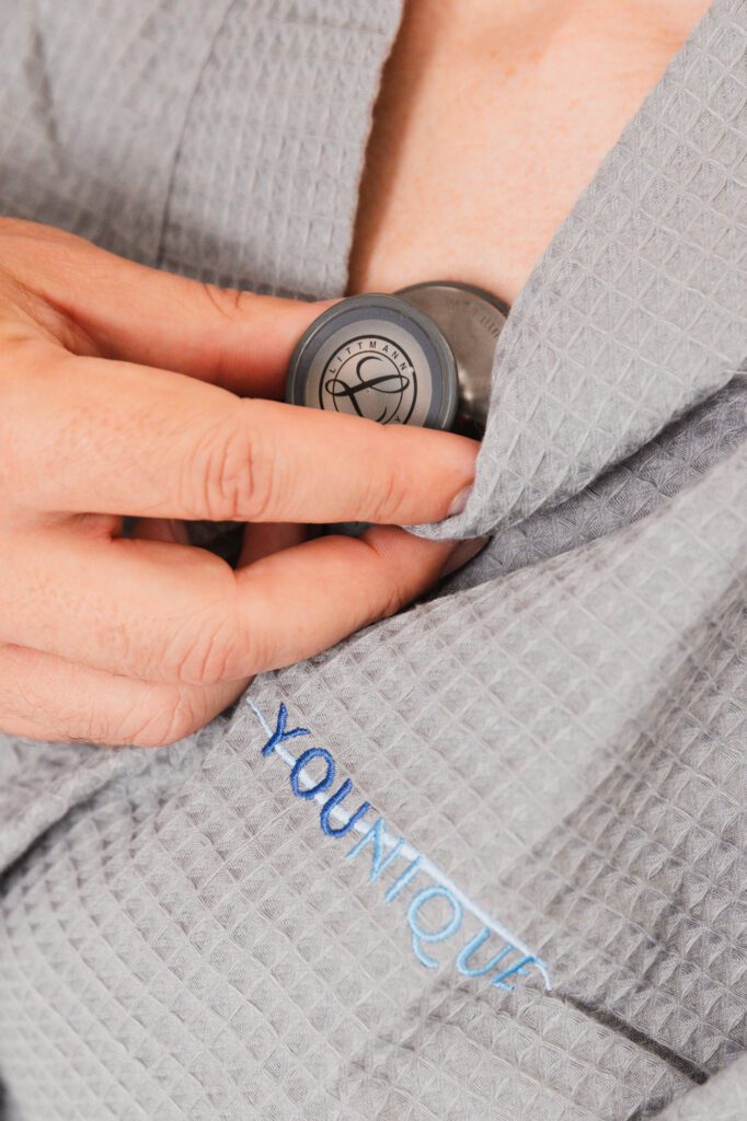 Person holding a stethoscope on chest wearing a gray robe with "Younique" embroidery.