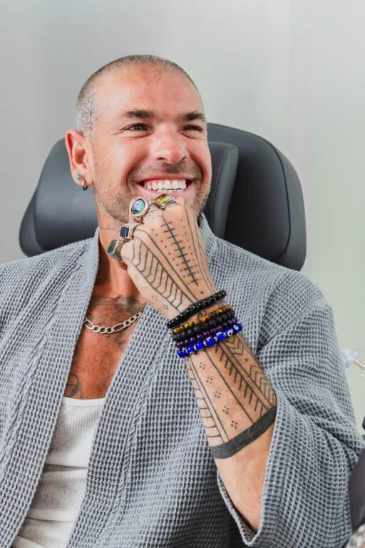 Smiling man with tattoos and rings, sitting in a clinic chair, showcasing cosmetic treatment satisfaction.