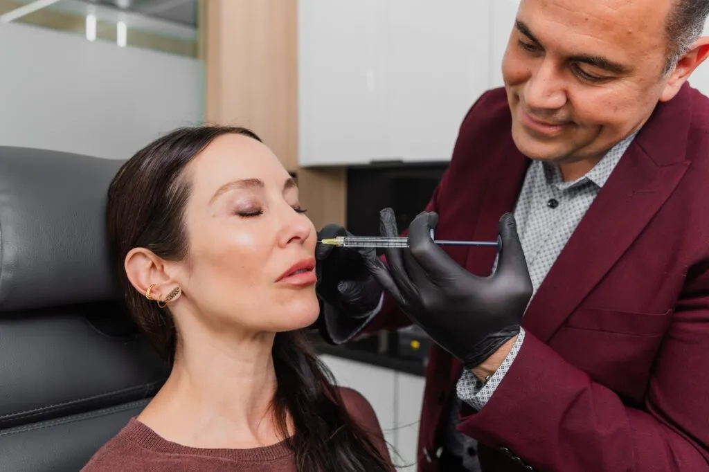Plastic surgeon performing a dermal filler injection on a woman's lips in a clinic setting.