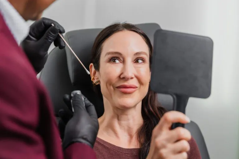 Woman at plastic surgery consultation holding a mirror while surgeon assists with a procedure.