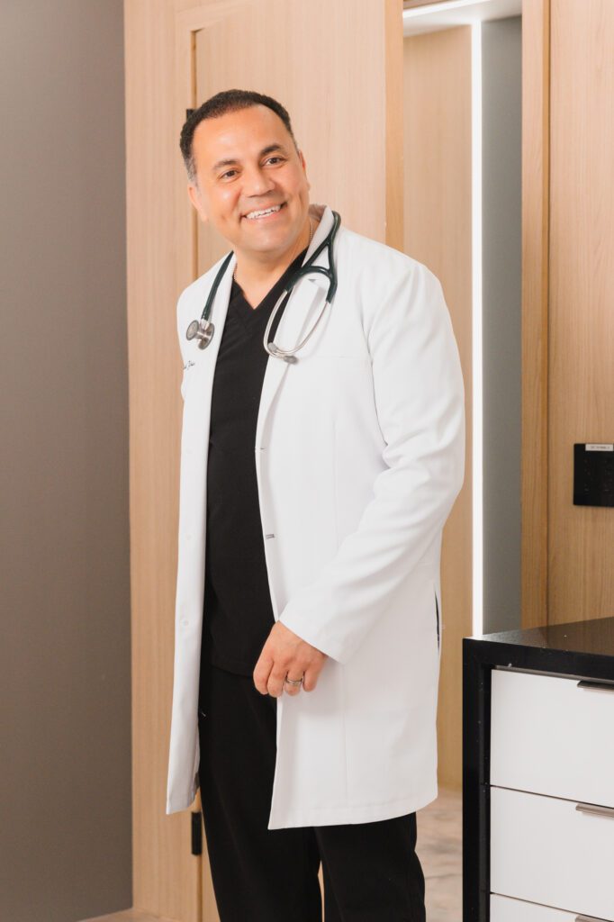 Plastic surgeon in a white coat with a stethoscope smiling in an office setting.