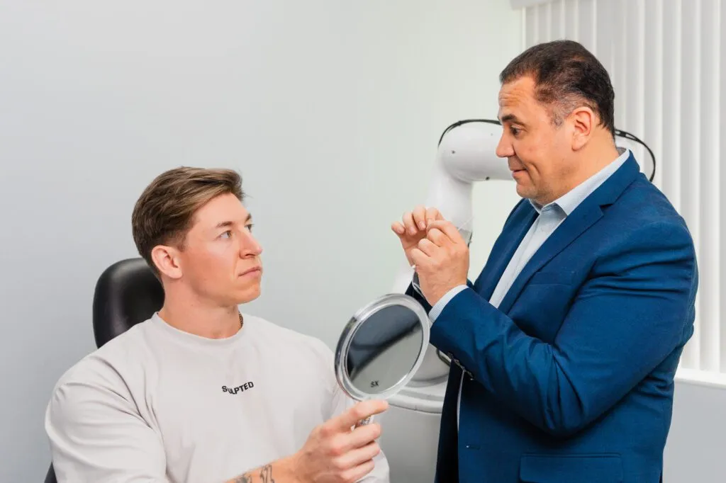 Plastic surgeon consulting male patient holding a mirror in a clinic setting.