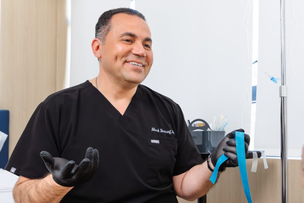 Smiling plastic surgeon in black scrubs holding medical tape in a clinic setting.