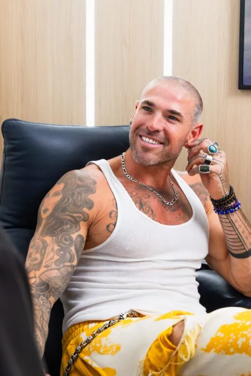Smiling person with tattoos and jewelry sitting on a chair at a plastic surgery center.