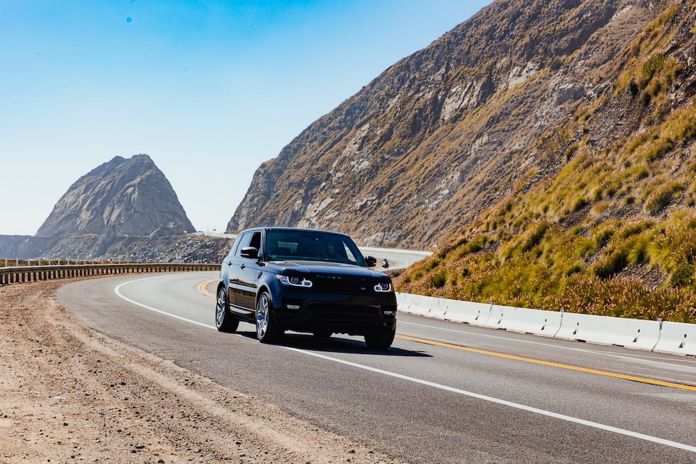 A sleek black SUV drives along a scenic coastal highway with mountains in the background.