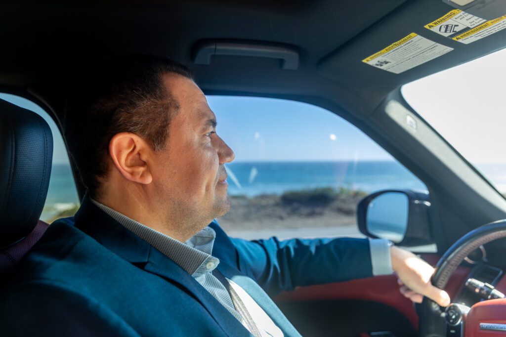 Man driving a car near the ocean on a sunny day, representing lifestyle and confidence.