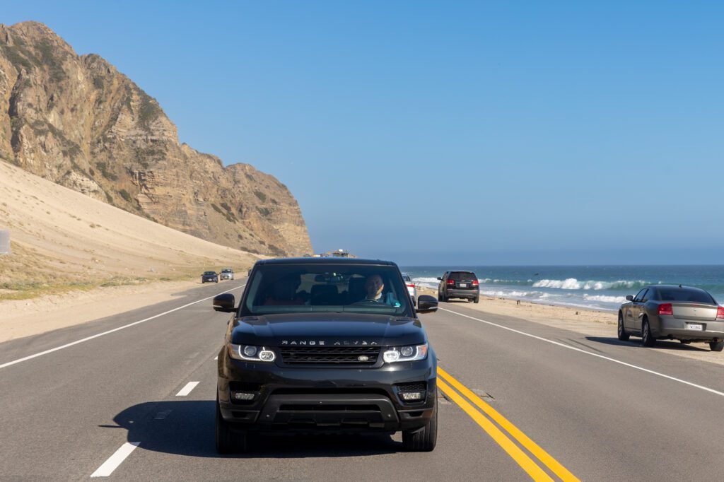 A black SUV drives along a scenic coastal highway with ocean and cliffs in the background.