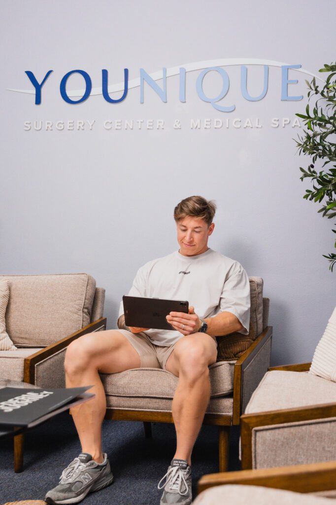 Man sitting in a modern waiting room of a plastic surgery center, reading a tablet.