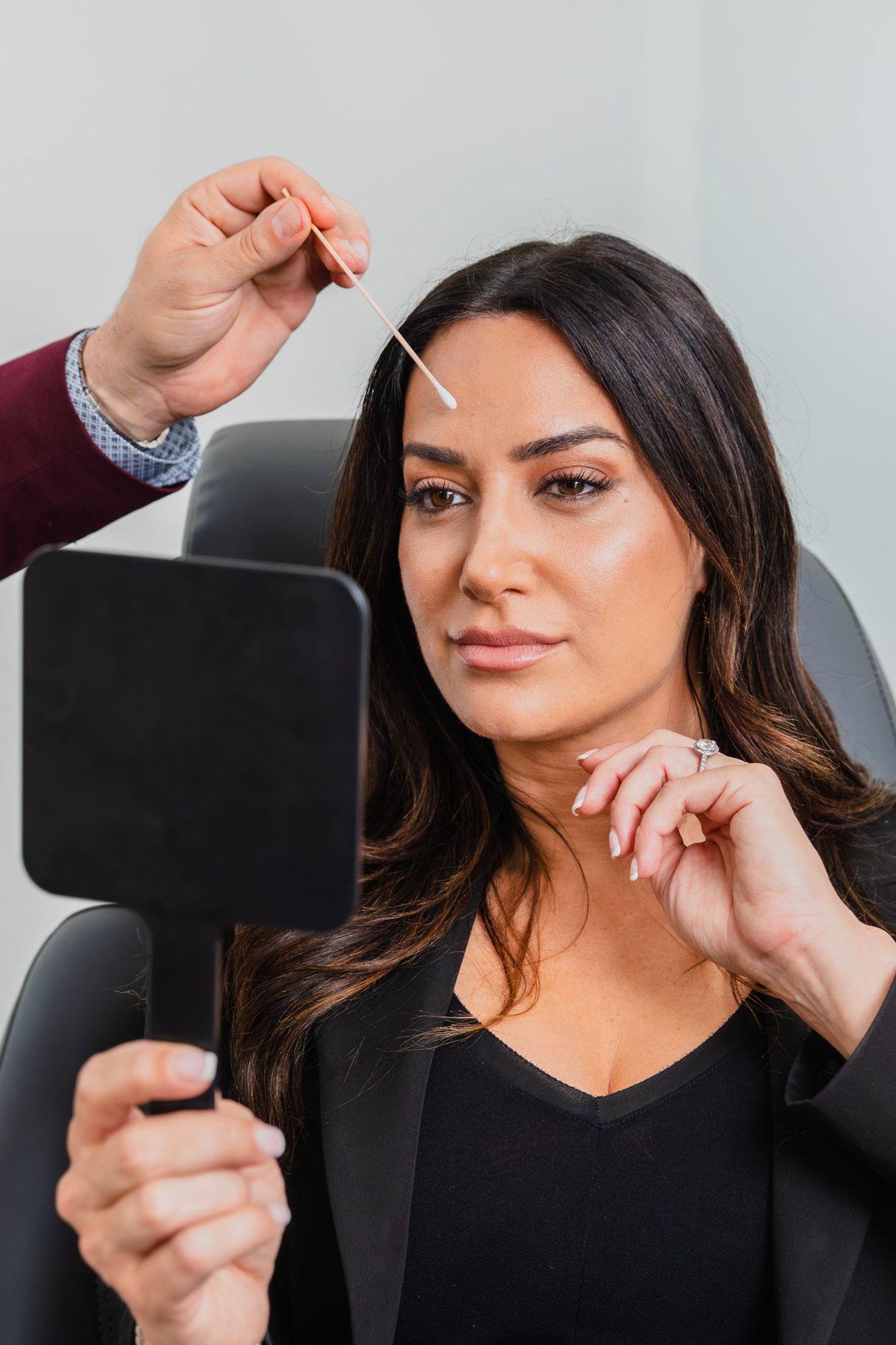 Woman contemplating cosmetic procedure while holding hand mirror, consultation at plastic surgery clinic.