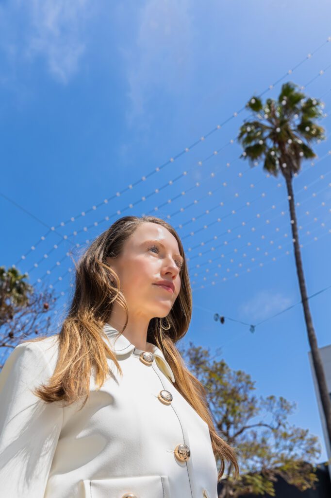 Woman in a white coat looking confident under a sunny blue sky.