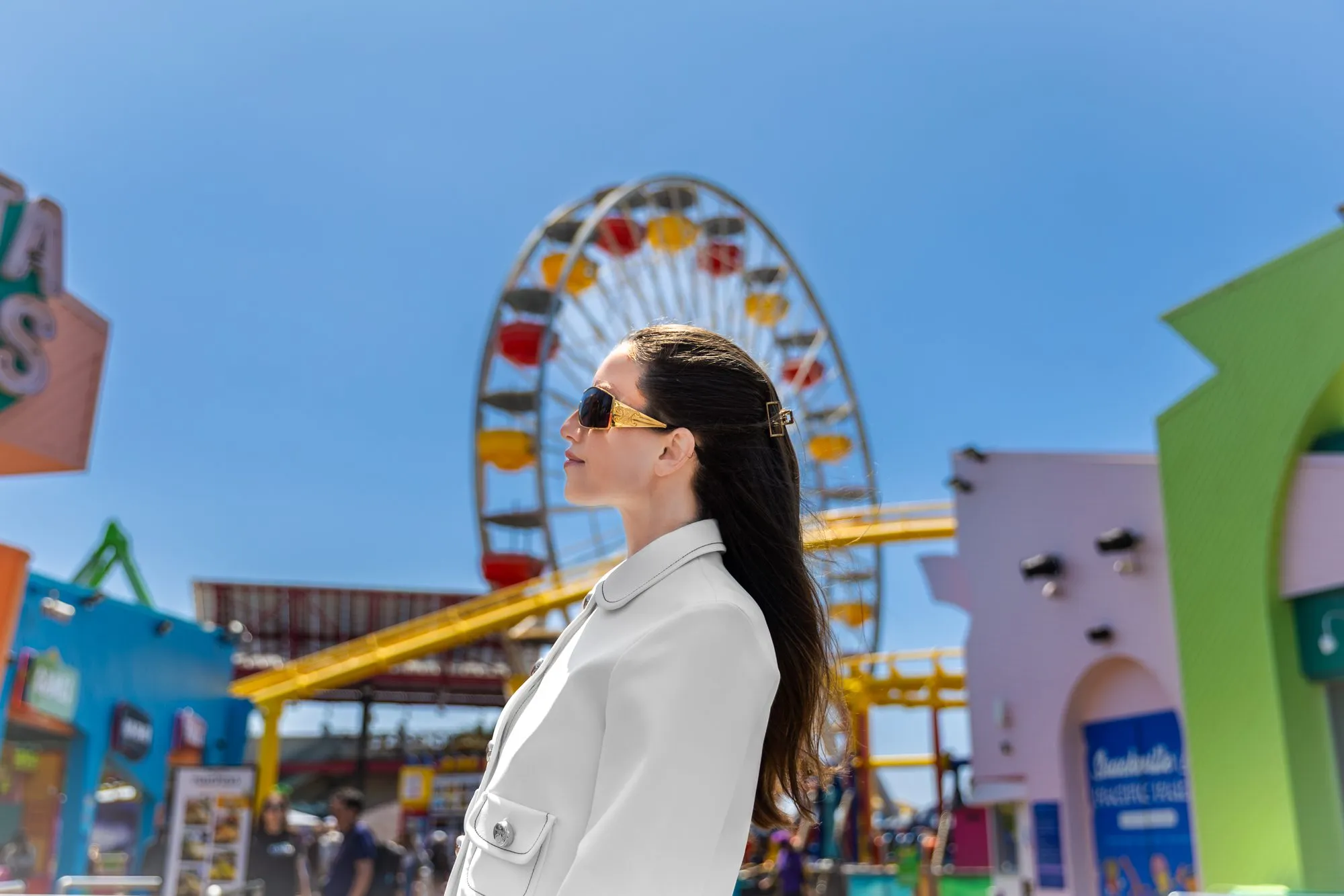 Woman in sunglasses and white coat at colorful amusement park with Ferris wheel in the background on a sunny day.