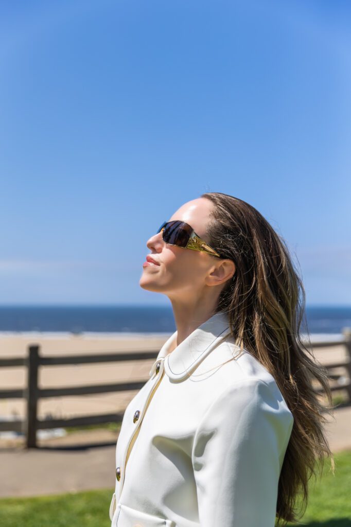 Woman in white coat with sunglasses smiling outdoors near beach.