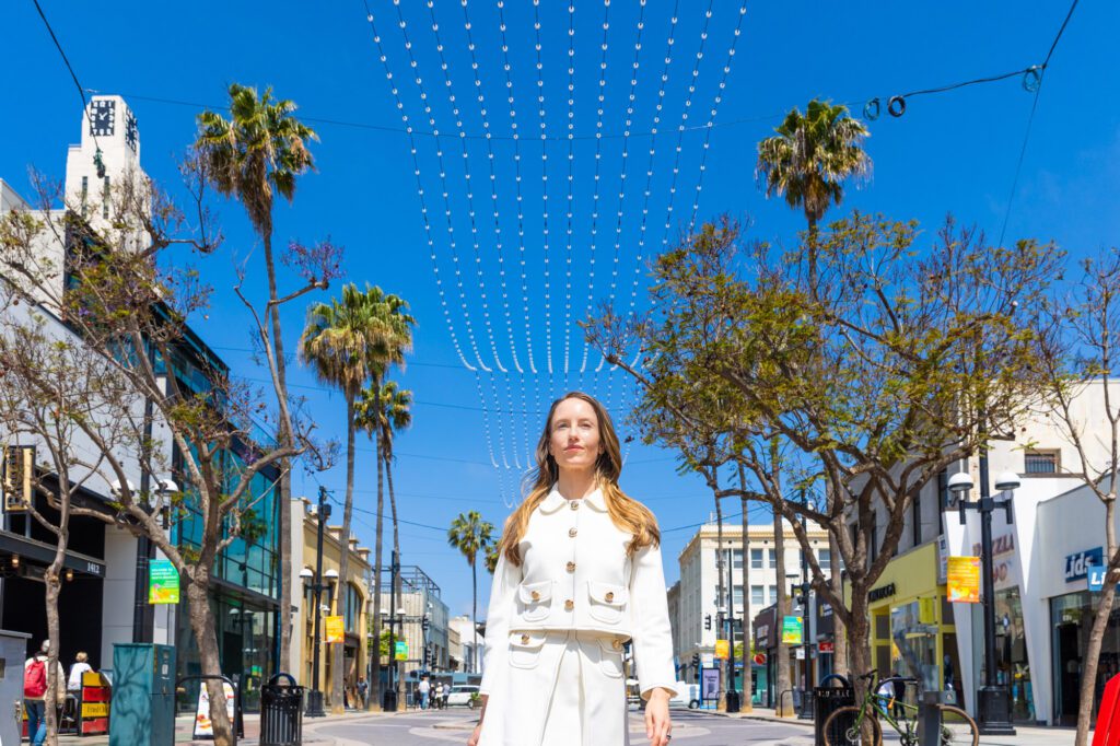 Woman in white coat walking confidently down a palm-lined street under a blue sky.