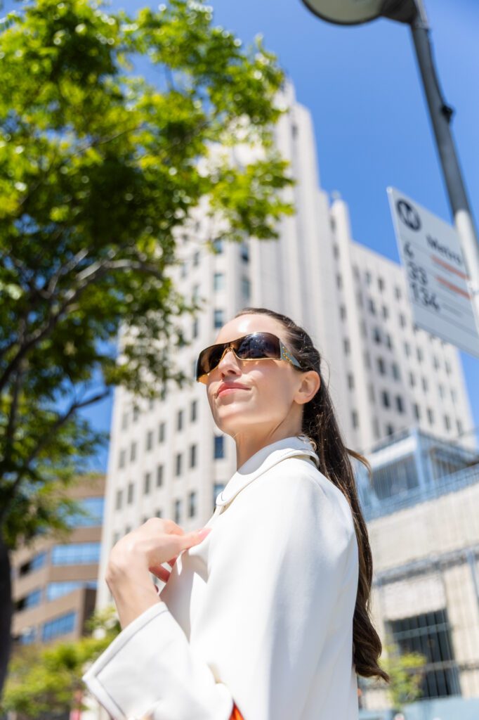 Woman in sunglasses and white coat confidently walking in a sunny urban setting.